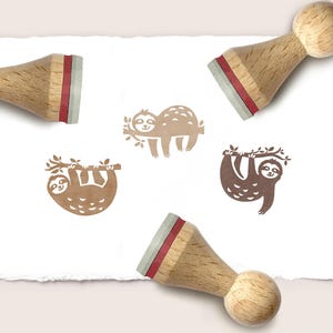 May include: Three wooden stamps with light-coloured handles and red rubber cushions. The stamps are positioned around three brown ink impressions on white paper, each depicting a sloth hanging from a tree branch in a different pose.