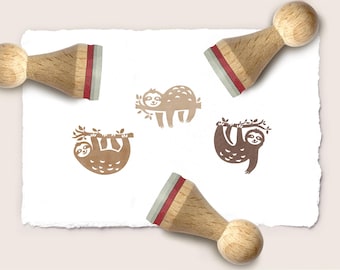 May include: Three wooden stamps with light-colored handles and red rubber cushions. The stamps are positioned around three brown ink impressions on white paper, each depicting a sloth hanging from a tree branch in a different pose.