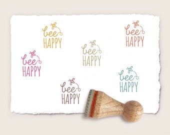 May include: A wooden stamp with the words "bee HAPPY" in a whimsical font, accompanied by a small bee graphic. The stamp has a wooden handle and a red and brown ink pad. Several impressions of the stamp are visible in various colors.