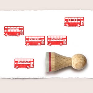 May include: A wooden rubber stamp with a round handle and a red rubber base, shown next to four red ink impressions of a double-decker bus on white paper. Each bus graphic features the word LONDON in white capital letters on the side.