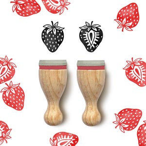 Può includere: Due timbri di fragole in legno con cuscinetti rossi e grigi. I timbri sono circondati da illustrazioni di fragole rosse su sfondo bianco. Sono presenti anche due illustrazioni di fragole in bianco e nero.