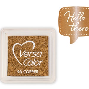 Könnte beinhalten: Ein goldfarbenes Stempelkissen mit einem Herz und dem Text "Versa Color" und "93 COPPER". Das Stempelkissen befindet sich in einem durchsichtigen Kunststoffgehäuse. Im Hintergrund befindet sich eine Sprechblase mit dem Text "Hello there".