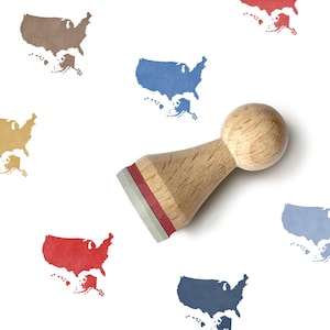 May include: A wooden stamp with a round handle and a rectangular base, featuring a red stripe. The stamp is next to multiple colorful United States map outlines in various colors, including blue, red, and brown. The background is white.
