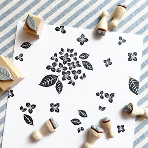 May include: A set of wooden stamps with various designs, including leaves and hydrangeas. The stamps are arranged on a white background with a blue and white striped fabric in the background.