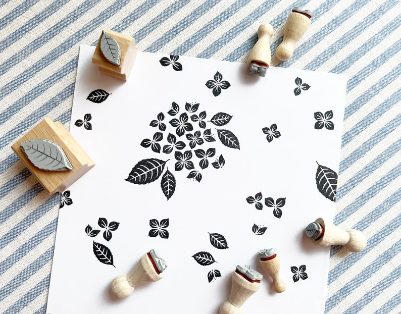 May include: A set of wooden stamps with various designs, including leaves and hydrangeas. The stamps are arranged on a white background with a blue and white striped fabric in the background.
