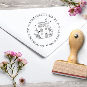 May include: A wooden address stamp featuring a house design and the text "SARA COLETTE ALBERT AM KIRCHBERG 181 83646 BAD TÖLZ". The stamp rests on a white envelope, with pink flowers.
