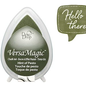 Könnte beinhalten: Ein grünes VersaMagic Kreide-Stempelkissen in einem tropfenförmigen Behälter mit dem Text "Hint of Pesto" und "Touche de pesto" und "Toque de pesto". Das Stempelkissen ist mit "Dew Drop" und "VersaMagic" beschriftet. Im Hintergrund befindet sich eine Sprechblase mit dem Text "Hello there".