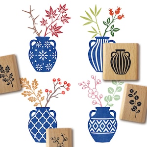 Op de afbeelding: Een verzameling houten stempels met botanische en vaasontwerpen, met stempelafdrukken van blauwe vazen met witte patronen, rode esdoornbladeren, groene bamboe, rode bessen en zwarte bloemtakjes op een witte achtergrond.