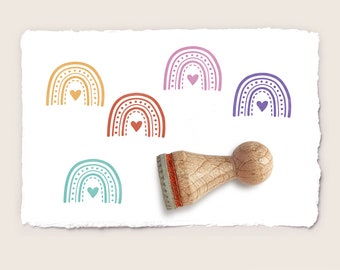 May include: A collection of colorful rainbow stamps with heart accents. The stamps are in shades of orange, red, teal, purple, and pink. A wooden stamp handle is visible on the right side. The stamps are on a white, textured paper.