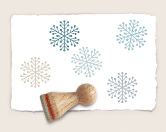 May include: A wooden stamp with a blue and beige snowflake design. The stamp is on a white piece of paper with four snowflake impressions.