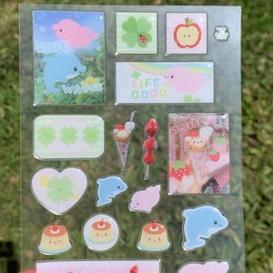 May include: A clear sticker sheet featuring various glittery designs including pink and blue dolphins, green clovers, red apples, puddings, and strawberry treats. Text includes "RIDE THE WAVES", "LIFE IS GOOD", "how SWEET!", and "so YUMMY".