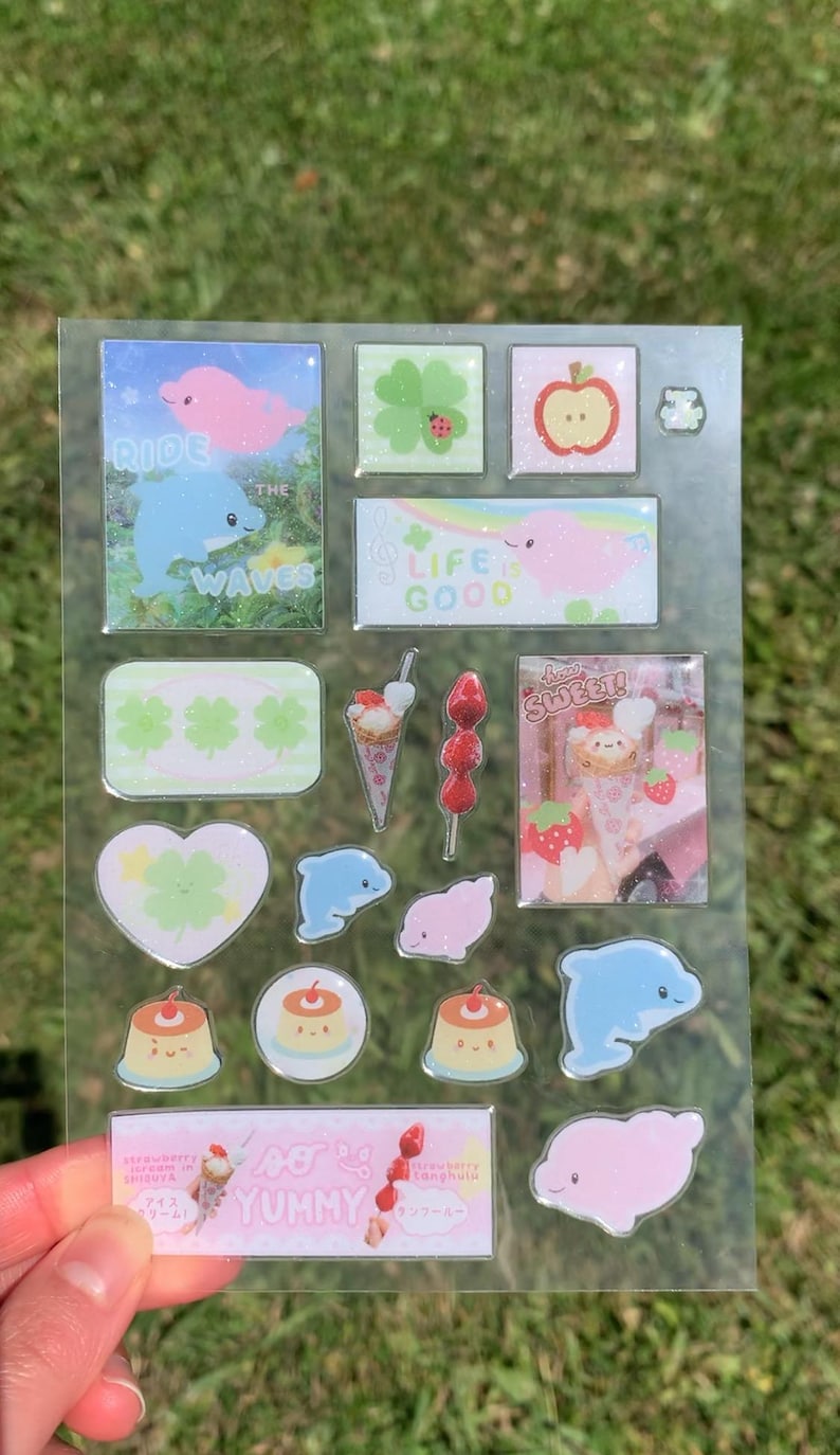 May include: A clear sticker sheet featuring various glittery designs including pink and blue dolphins, green clovers, red apples, puddings, and strawberry treats. Text includes "RIDE THE WAVES", "LIFE IS GOOD", "how SWEET!", and "so YUMMY".