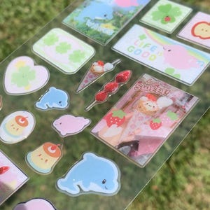 May include: A clear sticker sheet featuring various designs including blue dolphins, pink blobs, green clovers, a strawberry skewer, and rectangular scenes with text reading "WAVES," "LIFE IS GOOD," and "how SWEET!" alongside a cute ice cream cone graphic.