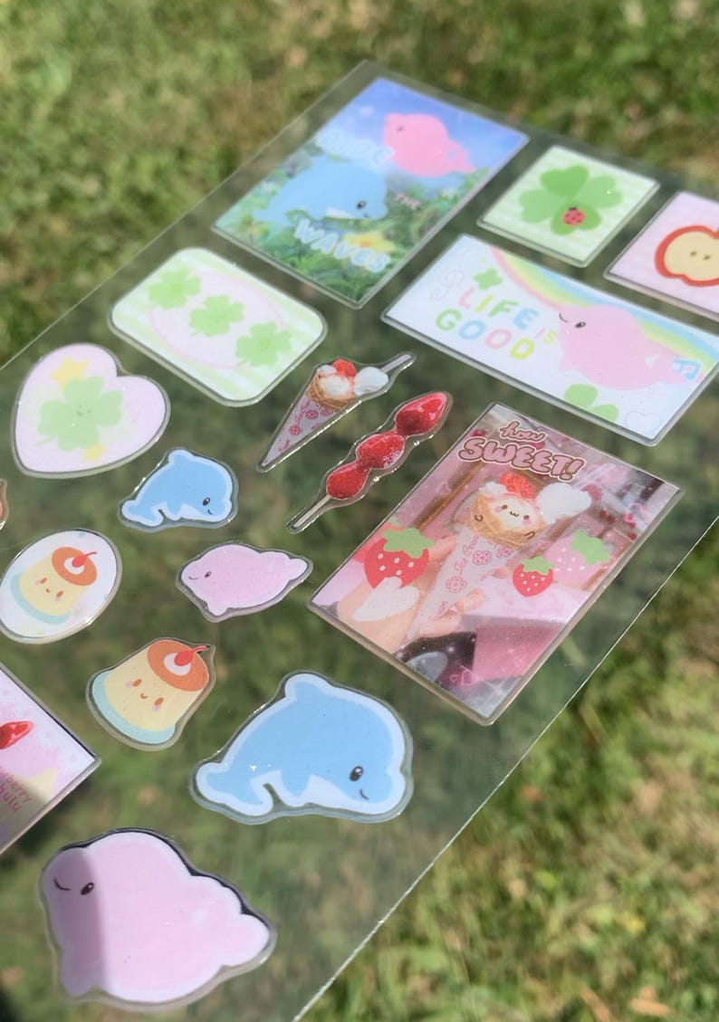 May include: A clear sticker sheet featuring various designs including blue dolphins, pink blobs, green clovers, a strawberry skewer, and rectangular scenes with text reading "WAVES," "LIFE IS GOOD," and "how SWEET!" alongside a cute ice cream cone graphic.
