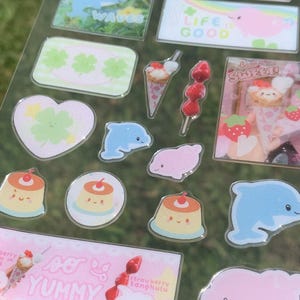 May include: A clear sticker sheet featuring various designs including blue dolphins, pink sea creatures, green clovers, red apples, custard puddings, strawberry tanghulu, and ice cream cones with text including "RIDE THE WAVES", "LIFE IS GOOD", "HOW SWEET!", and "SO YUMMY".