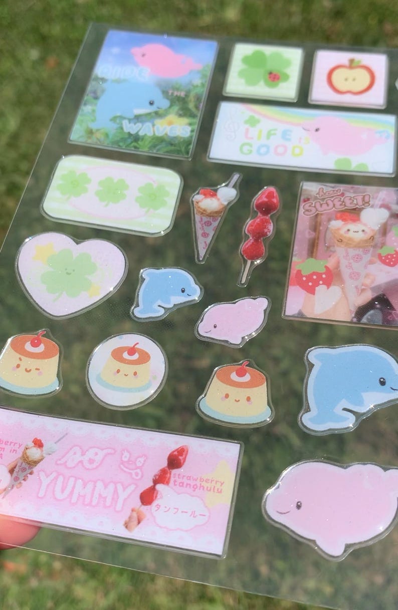 May include: A clear sticker sheet featuring various designs including blue dolphins, pink sea creatures, green clovers, red apples, custard puddings, strawberry tanghulu, and ice cream cones with text including "RIDE THE WAVES", "LIFE IS GOOD", "HOW SWEET!", and "SO YUMMY".
