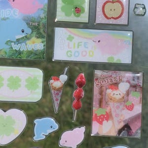 May include: A sticker sheet featuring various designs including a blue dolphin with the text "WAVES", a green clover with a ladybug, a red apple, a rectangular sticker with a pink creature and the text "LIFE IS GOOD", ice cream cones, strawberries, and puddings with the text "how SWEET!".