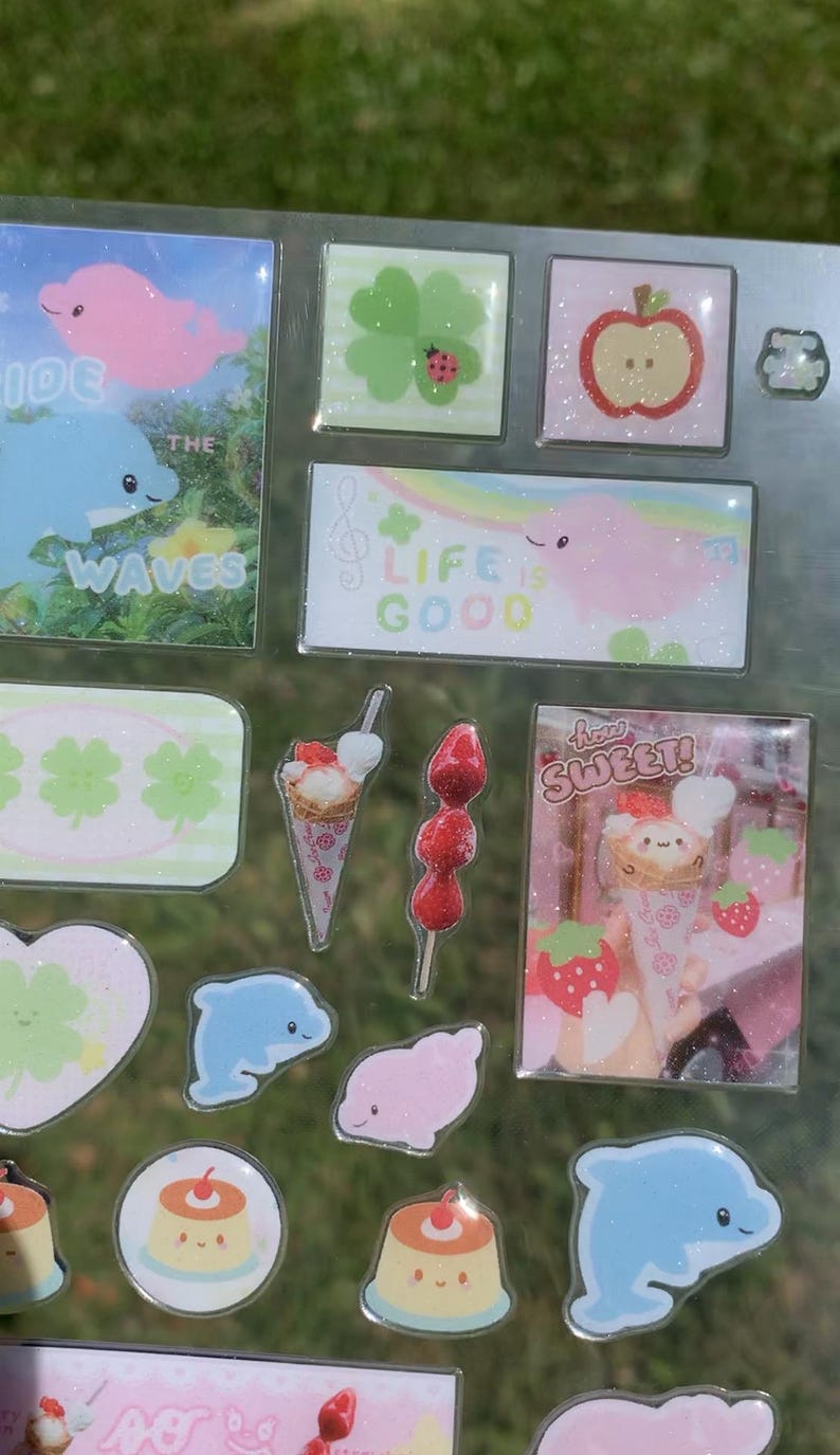 May include: A sticker sheet featuring various designs including a blue dolphin with the text "WAVES", a green clover with a ladybug, a red apple, a rectangular sticker with a pink creature and the text "LIFE IS GOOD", ice cream cones, strawberries, and puddings with the text "how SWEET!".