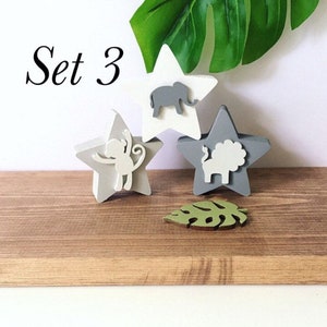 Op de afbeelding: Set van drie houten sterornamenten met dierensilhouetten. De sterren zijn geschilderd in grijstinten en wit. De dieren zijn een olifant, een aap en een leeuw. De set bevat ook een groen blaadornament. De tekst "Set 3" is zichtbaar in de afbeelding.