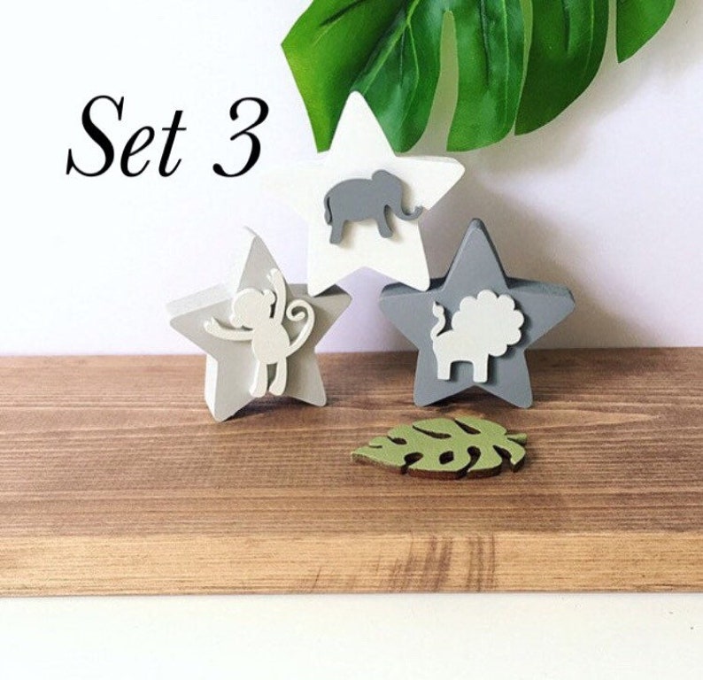 Op de afbeelding: Set van drie houten sterornamenten met dierensilhouetten. De sterren zijn geschilderd in grijstinten en wit. De dieren zijn een olifant, een aap en een leeuw. De set bevat ook een groen blaadornament. De tekst "Set 3" is zichtbaar in de afbeelding.