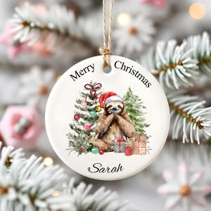 May include: A white ceramic Christmas bauble featuring a sloth in a Santa hat, with Christmas trees and gifts. The bauble has the words "Merry Christmas" and the name "Sarah" printed on it, and is hung by a natural twine.