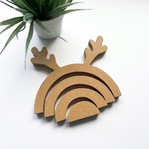 May include: A light brown, flat-surfaced wooden cutout shaped like a rainbow with three concentric arches. The top arch features two protruding, branched antler-like shapes extending upwards from the outer curve of the rainbow structure.