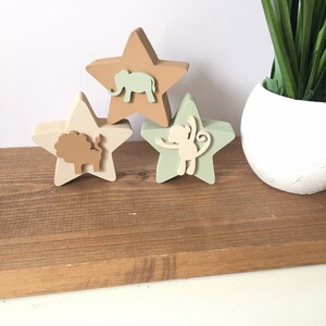 Op de afbeelding: Drie houten stervormige ornamenten met dierensilhouetten. De bovenste ster is bruin met een lichtbruine olifant, de middelste ster is cr&egrave;me met een bruine leeuw en de onderste ster is groen met een lichtbruine aap.