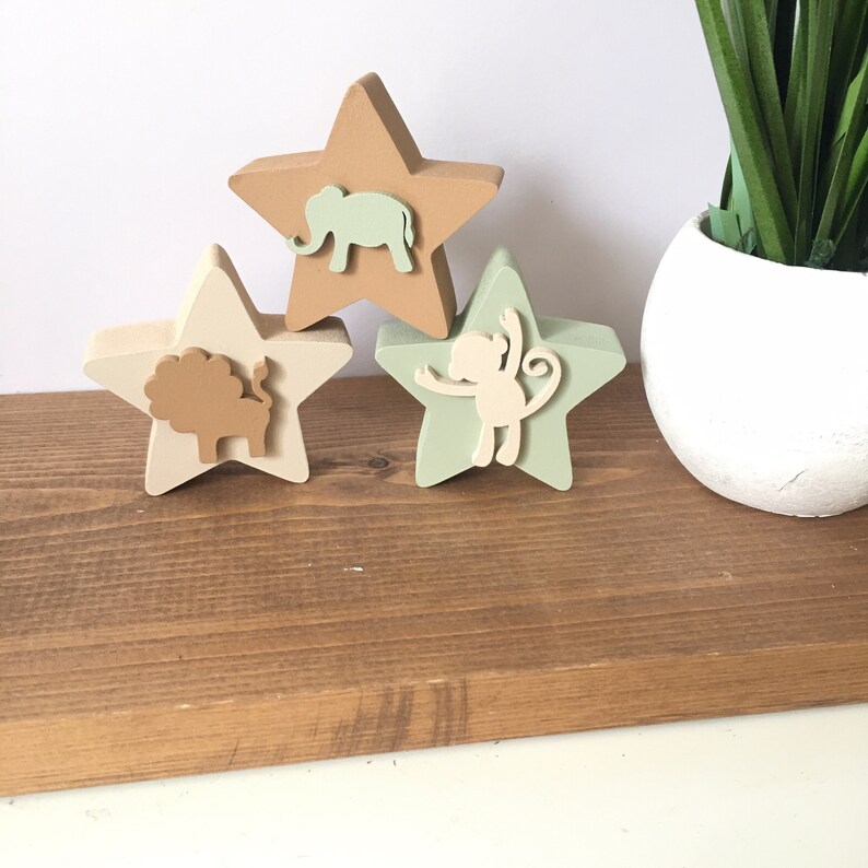 Op de afbeelding: Drie houten stervormige ornamenten met dierensilhouetten. De bovenste ster is bruin met een lichtbruine olifant, de middelste ster is cr&egrave;me met een bruine leeuw en de onderste ster is groen met een lichtbruine aap.