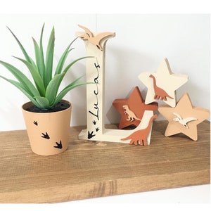 May include: Wooden letter L with the name "Lucas" written on it. The letter is decorated with dinosaurs and stars in shades of brown, orange, and cream. A small potted succulent plant with brown dinosaur footprints on the pot is in the foreground.