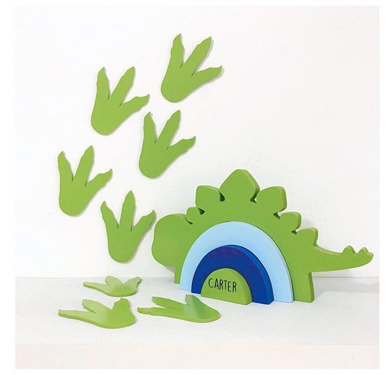 May include: Green wooden dinosaur cutout with a blue, light blue, and dark blue semi-circle base. The base has the name "CARTER" written on it. There are several green dinosaur footprints scattered around the dinosaur.