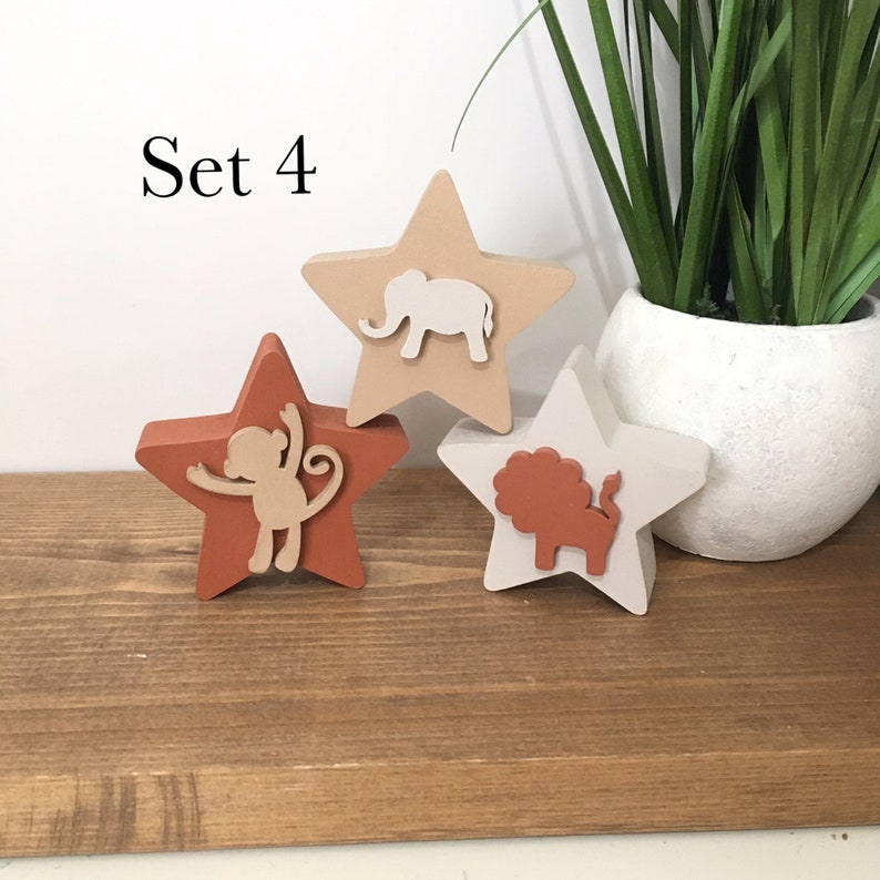 Op de afbeelding: Set van drie houten stervormige ornamenten met dierensilhouetten. De sterren zijn geschilderd in tinten bruin, beige en wit. De dieren zijn een olifant, een aap en een leeuw.