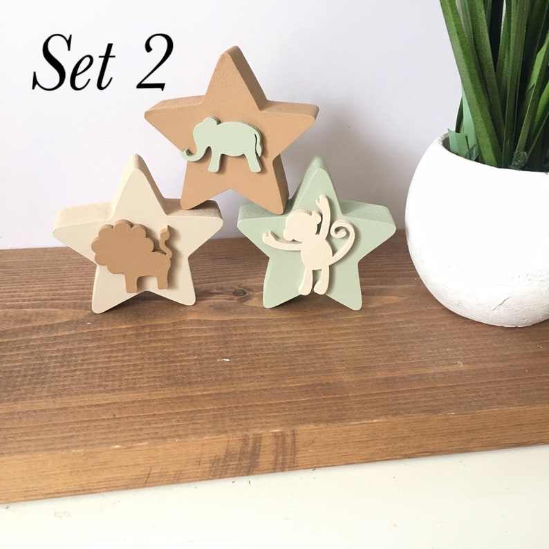 Op de afbeelding: Set van drie houten stervormige ornamenten met dierensilhouetten. De sterren zijn geschilderd in tinten bruin, beige en groen. De dieren zijn een olifant, een leeuw en een aap.