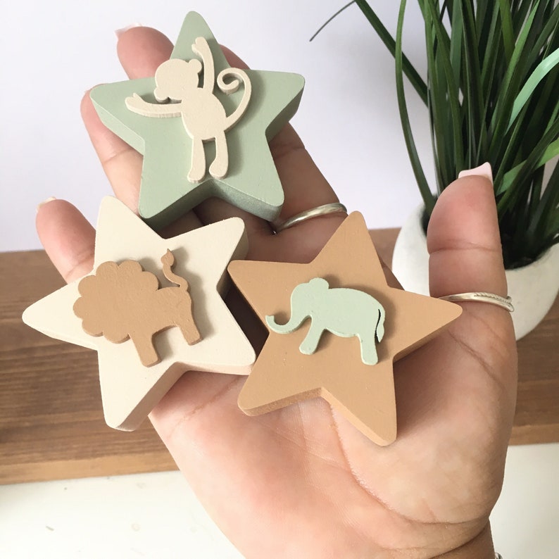 Op de afbeelding: Drie houten stervormige ornamenten met dierensilhouetten. De sterren zijn geschilderd in tinten groen, bruin en beige. De dieren zijn een aap, een leeuw en een olifant.