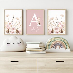 May include: Three framed floral prints and decorative items on a wooden surface. The prints feature botanical illustrations in soft pinks and browns. A cloud-shaped night light, books, and a rainbow decor piece complete the scene.