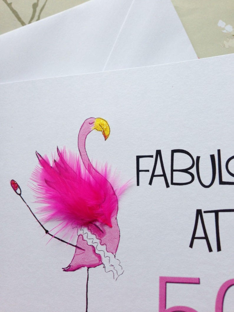 May include: A pink flamingo with a yellow beak and pink feathers is drawn on a white card. The flamingo is wearing a white tutu and has a pink feather on its head. The card says "Fabulous at 50".
