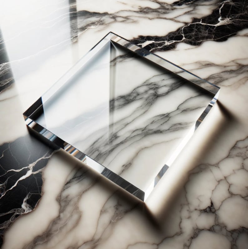 May include: A clear, square glass block with beveled edges. The glass reflects the light and the marble surface beneath it. The marble has a white and gray veined pattern.