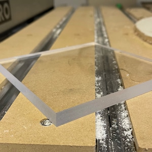 May include: A clear, square acrylic sheet with frosted edges. The sheet is positioned on a light brown surface, likely a workbench, with visible grooves and a small water droplet. The edges of the acrylic are smooth and reflect light.