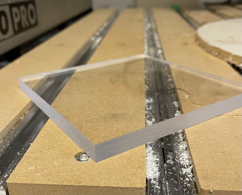 May include: A clear, square acrylic sheet with frosted edges. The sheet is positioned on a light brown surface, likely a workbench, with visible grooves and a small water droplet. The edges of the acrylic are smooth and reflect light.