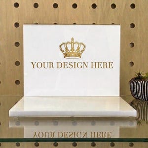 May include: A rectangular white acrylic sign holder with a horizontal base. The front panel features a gold-colored graphic of a crown centered above the text "YOUR DESIGN HERE" in a gold-colored serif font. The sign is clear and reflective.