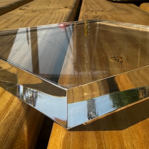 May include: A clear, square glass object with beveled edges. The glass reflects the surrounding environment, creating a mirrored effect. The object is positioned on a wooden surface, with the grain of the wood visible.
