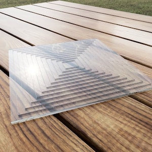 May include: A clear glass square with a concentric square pattern. The glass is sitting on a wooden surface.
