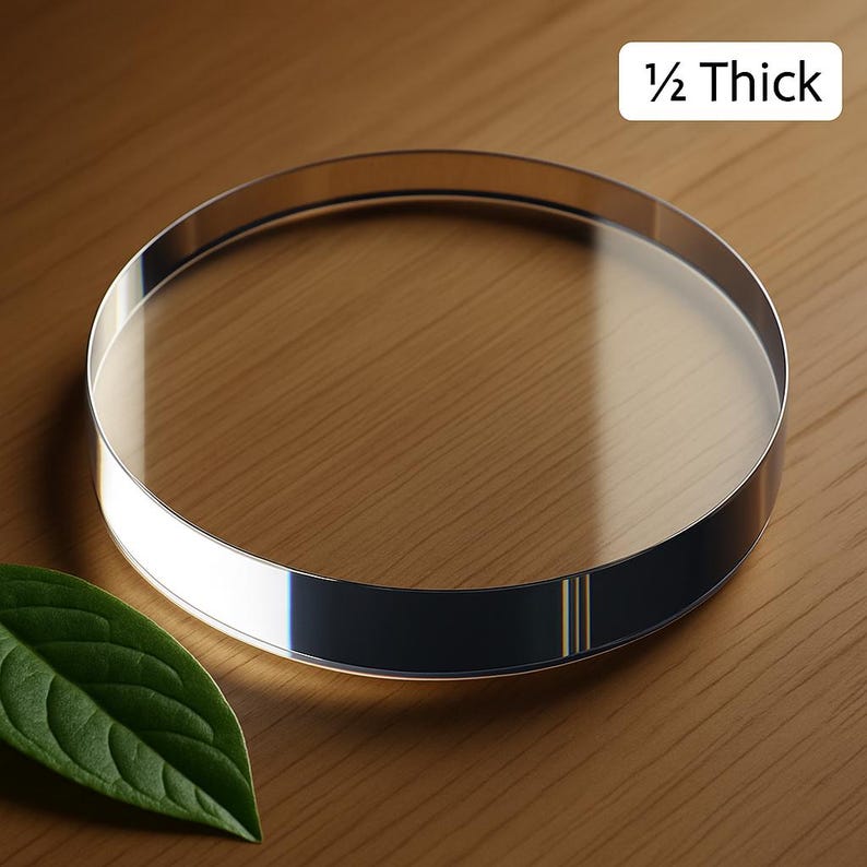 May include: A clear, circular glass object with a polished silver band. The text "1/2 Thick" is in the upper right corner. The object is on a wooden surface, with a green leaf in the lower left corner.