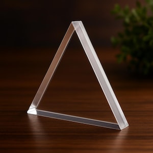 May include: A clear, transparent acrylic triangle with a flat base. The geometric shape is displayed on a dark wooden surface, with a blurred green plant in the background. The edges of the triangle are smooth and polished.