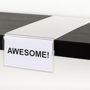 May include: A clear acrylic shelf talker sign holder with a rectangular design that folds over the edge of a shelf. The holder contains a white paper insert with the word AWESOME! printed in bold black capital letters on the front face.