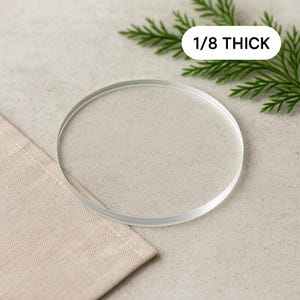 May include: A clear, circular coaster, 1/8 inch thick, sits on a beige linen napkin. The coaster is transparent and has a smooth surface. Green sprigs are in the background. The text "1/8 THICK" is in a white rectangle.