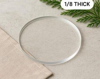 May include: A clear, circular coaster, 1/8 inch thick, sits on a beige linen napkin. The coaster is transparent and has a smooth surface. Green sprigs are in the background. The text "1/8 THICK" is in a white rectangle.