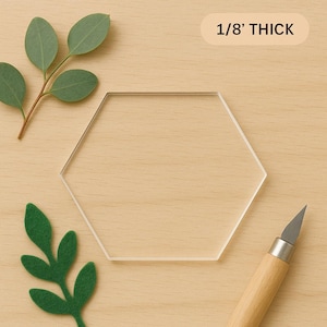 May include: A clear, hexagonal acrylic piece, 1/8" thick, sits on a light wood surface. Green felt leaves and a sprig of eucalyptus are arranged around the hexagon. A craft knife with a wooden handle is in the lower right corner.