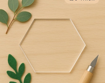 May include: A clear, hexagonal acrylic piece, 1/8" thick, sits on a light wood surface. Green felt leaves and a sprig of eucalyptus are arranged around the hexagon. A craft knife with a wooden handle is in the lower right corner.