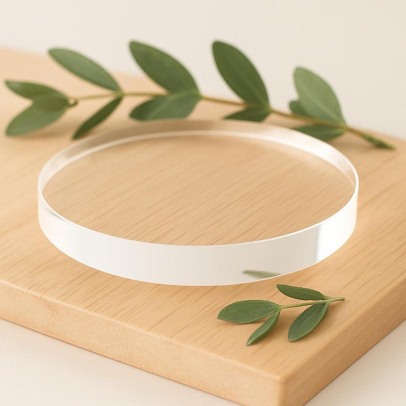 May include: A clear, circular acrylic display stand with a white edge sits on a light brown wooden surface. Green leaves are arranged around the stand. The stand is approximately 8 inches in diameter.