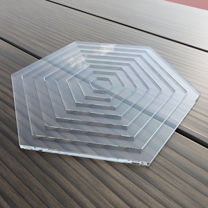 May include: A clear acrylic hexagon with five concentric rings, creating a layered effect. The hexagon is resting on a wooden surface.